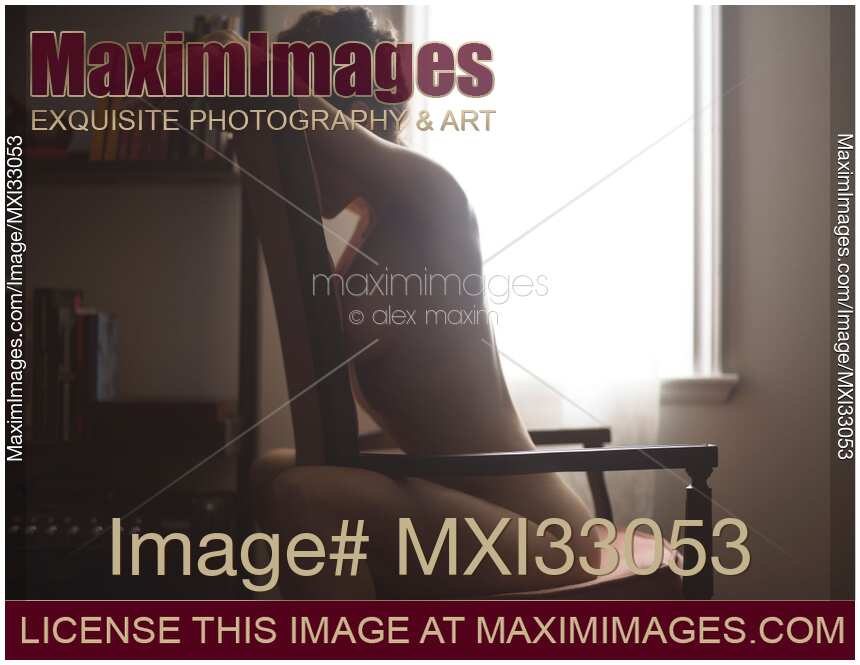 Artistic nude portrait of a beautiful woman sitting naked facing armchair backrest by a window