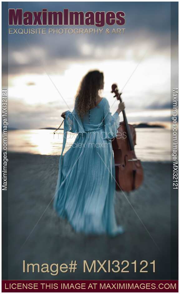 Atmospheric portrait of a woman in a vintage dress standing with a cello on an ocean shore in the wind in dramatic sunset