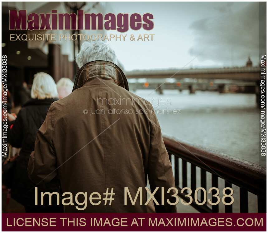 Back of a grey-haired man in winter jacket walking along the river in London moody city scenery