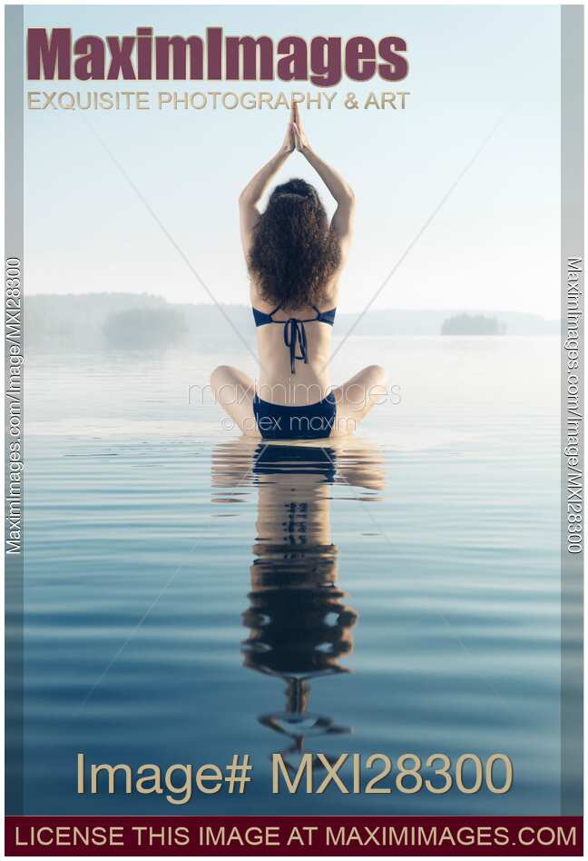 Back of a woman practicing yoga meditation on the water in sunrise mist