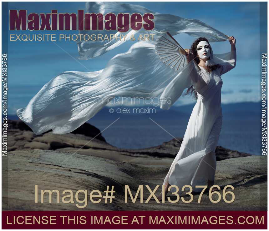 Beautiful asian woman in mysterious white mask performing dramatic graceful dance with silk fans in the wind on seashore