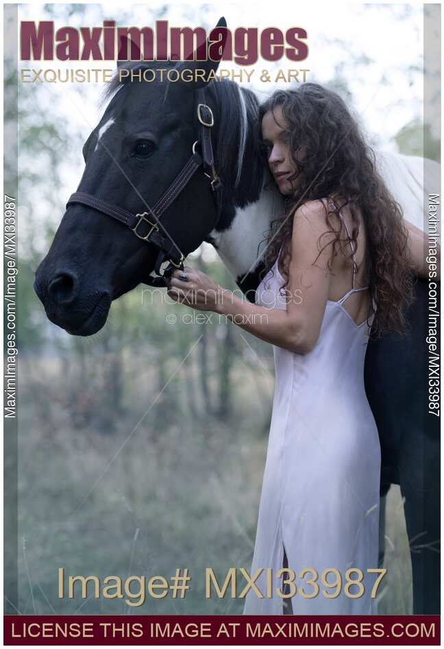 Beautiful girl with long curly dark hair hugging a black and white horse. Expressive candid portrait.