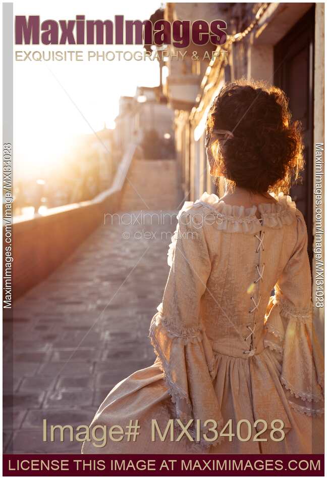 Beautiful lady in vintage Venetian carnival dress and mask standing on sunlit historic street in Venice