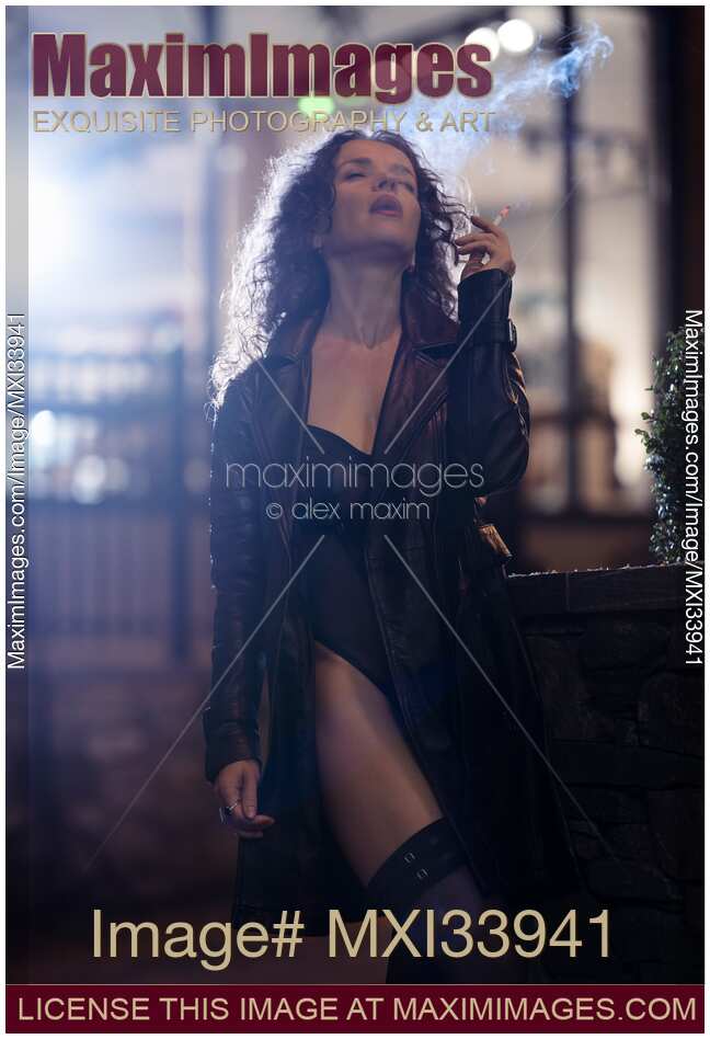 Beautiful seductive woman in black leather coat and sexy lingerie smoking cigarette on night city street