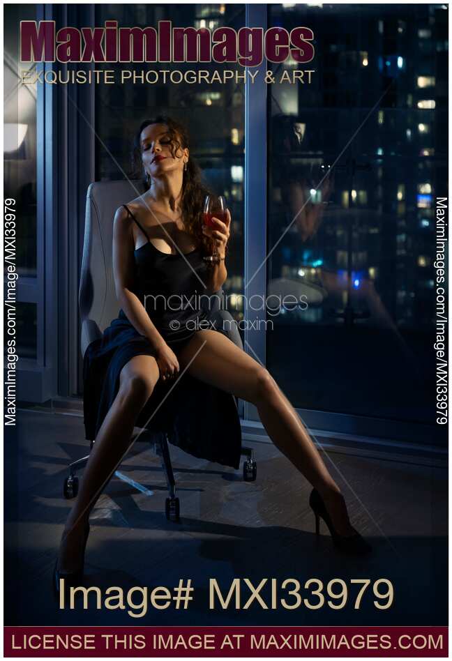 Beautiful seductive woman with glass of wine sitting in chair in provocative pose by window with night city view