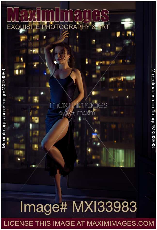 Beautiful sexy woman in black dress standing in a provocative pose baring her leg by a condo window with night city view