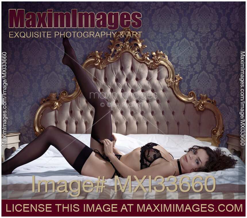 Beautiful sexy woman in black lingerie putting on stockings on her legs lying on antique bed with a provocative expression
