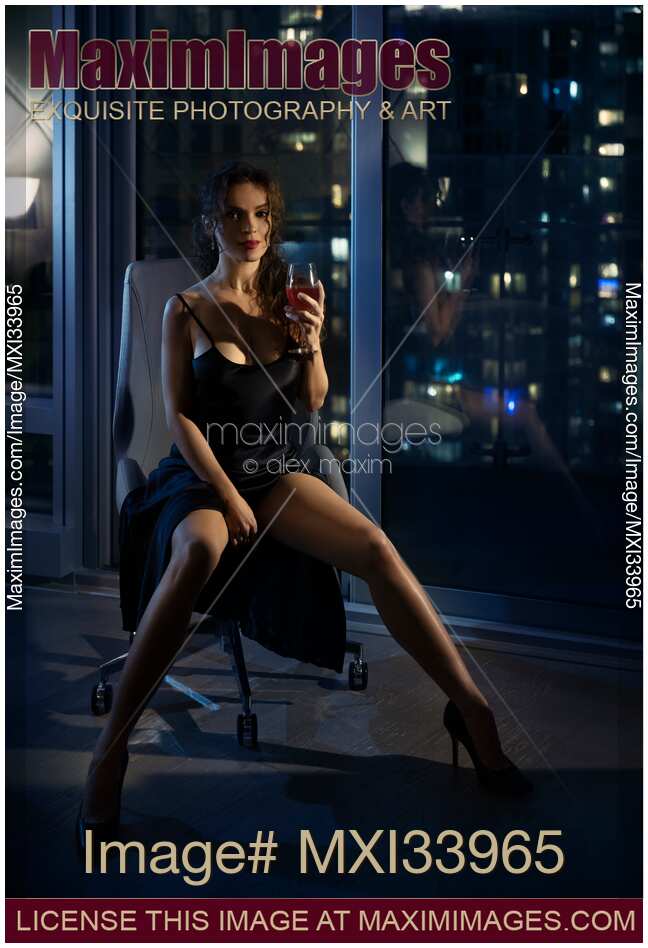Beautiful sexy woman with glass of wine sitting in chair in provocative pose by a condo window at night