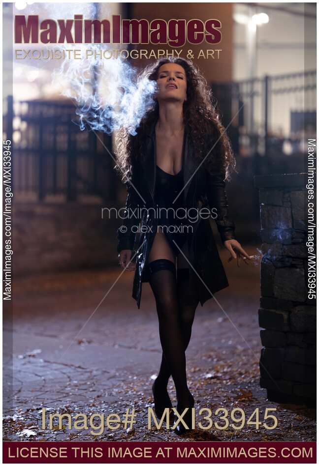Beautiful woman in sexy provocative black clothes and stockings exhaling cigarette smoke with playful expression on night city street