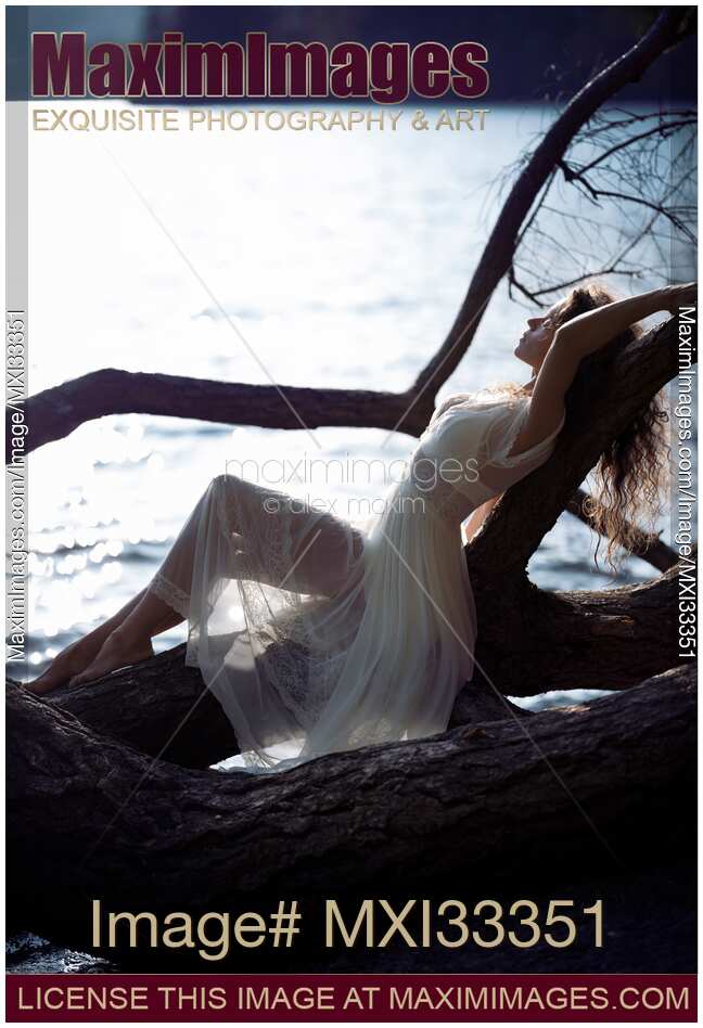 Beautiful woman in sheer white lace dress reclining relaxed against a tree by the lake in the nature