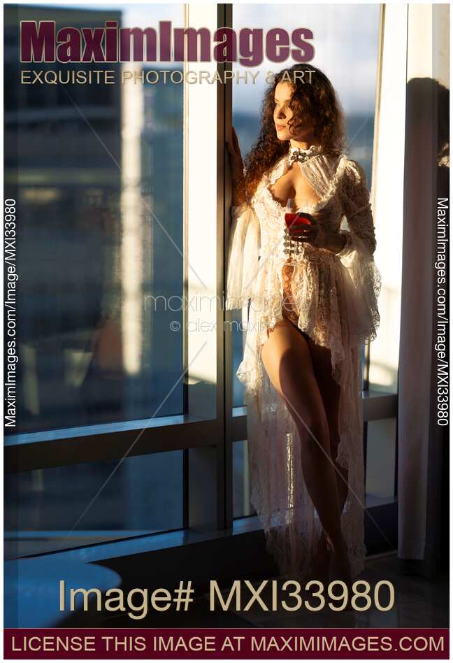 Beautiful woman in white lace night gown standing half naked with a glass of wine by a window in sunset light