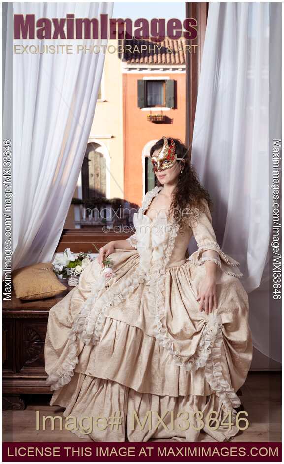 Beautiful young lady in vintage Italian dress and carnival mask sitting by an open window in Venice