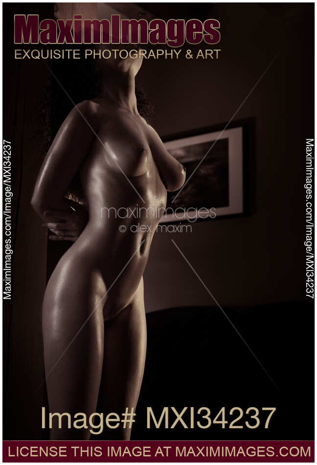 Black and white art nude photo of a beautiful naked woman with shiny skin leaning against a wooden pillar in a dark room