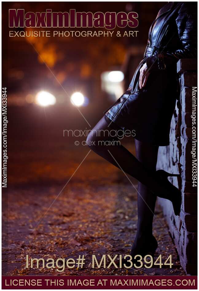 Closeup of sexy woman in black leather coat, stockings and high heel shoes standing on night city street in car lights