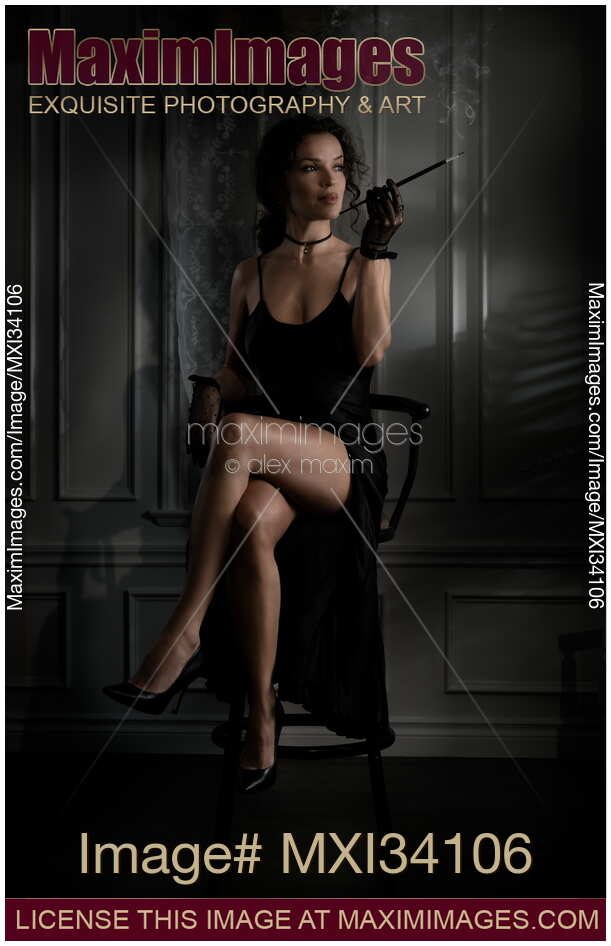 Elegant lady in black dress revealing her sexy legs sitting holding long cigarette holder in dark room