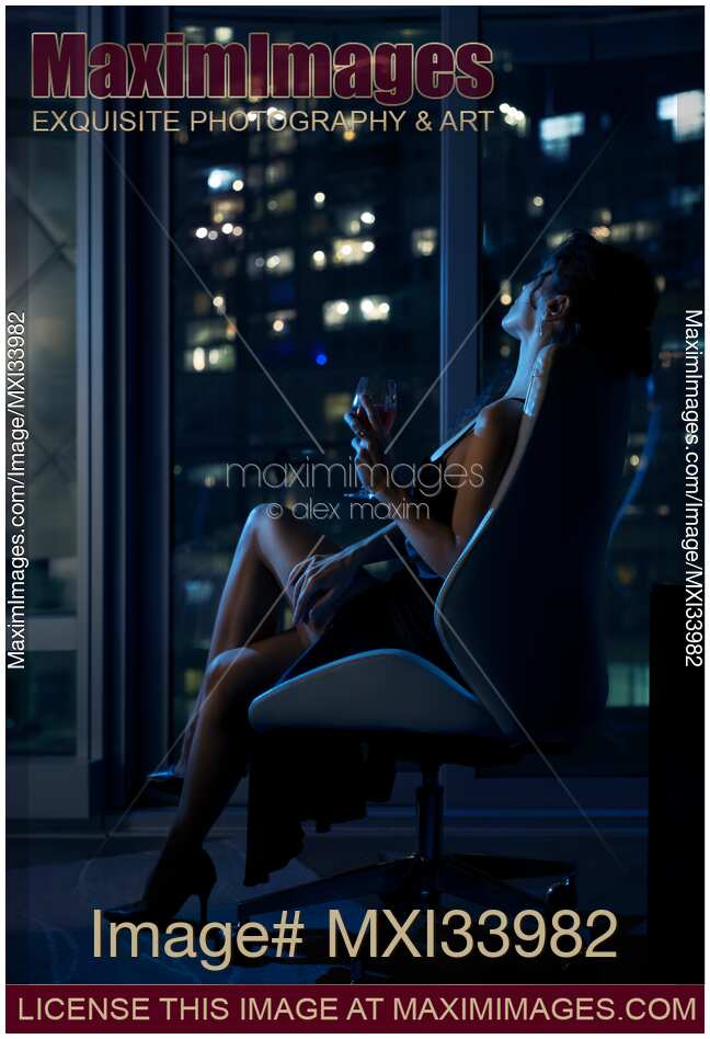 Elegant woman sitting relaxed in chair with glass of wine by window with night city view