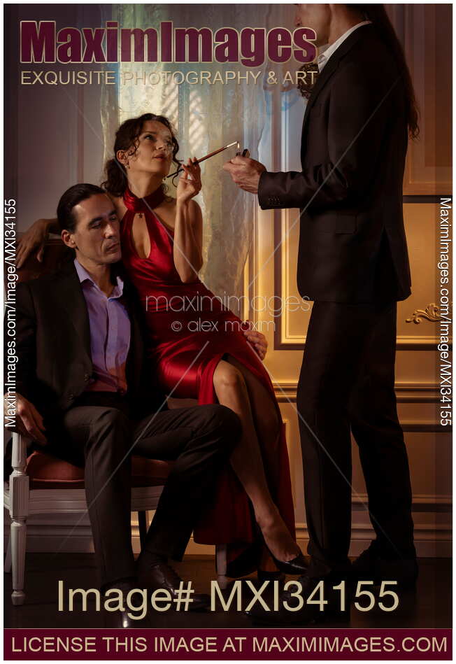 Gentleman lights a cigarette of a beautiful lady in red evening dress sitting with another man. Suggestive romantic trio.