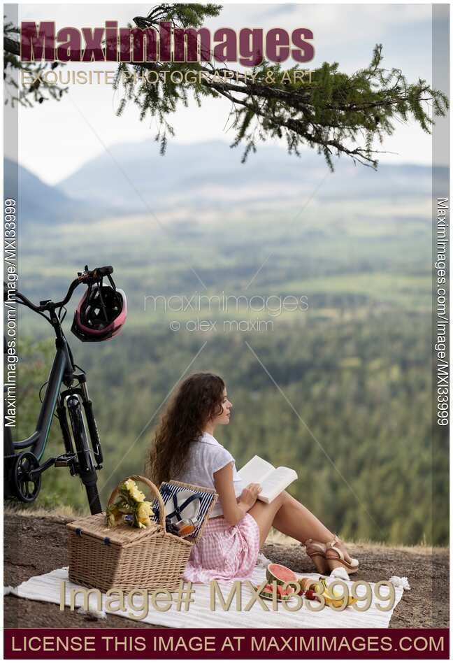 Girl sitting on a blanket with a book and a bike enjoying a little picnic in summer nature scenery