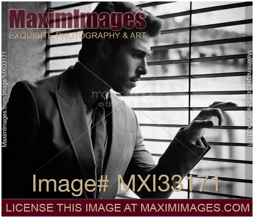 Handsome young man in classic gray suit looking out through window blinds with concerned expression