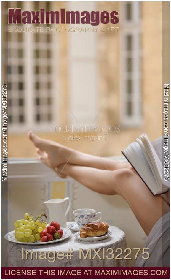 Legs of a woman reading a book by open window with French breakfast of croissant and coffee