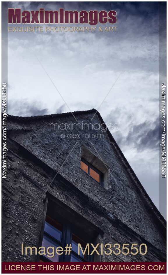 Lit up gable wall attic window of an old house at night under dramatic cloudy sky