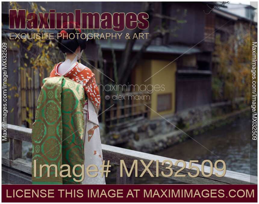 Maiko young Geisha apprentice in a red kimono with long obi standing on a bridge