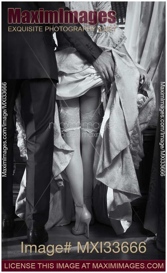 Man raising up woman's dress skirt revealing her sexy legs in stockings and high heels. Black and white rear view.