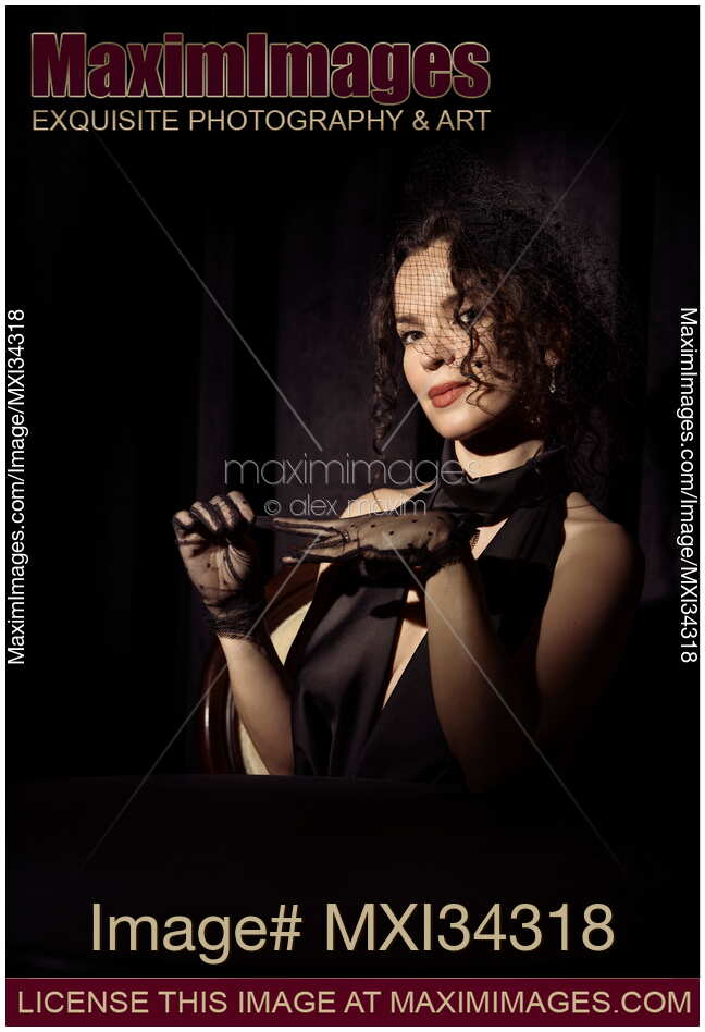 Mysterious femme fatale lady in black wearing veiled hat taking off her gloves with mischievous smile