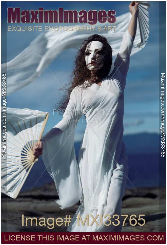 Mysterious ghost-like asian woman in white face mask and flowy dress dancing with silk hand fans in the wind