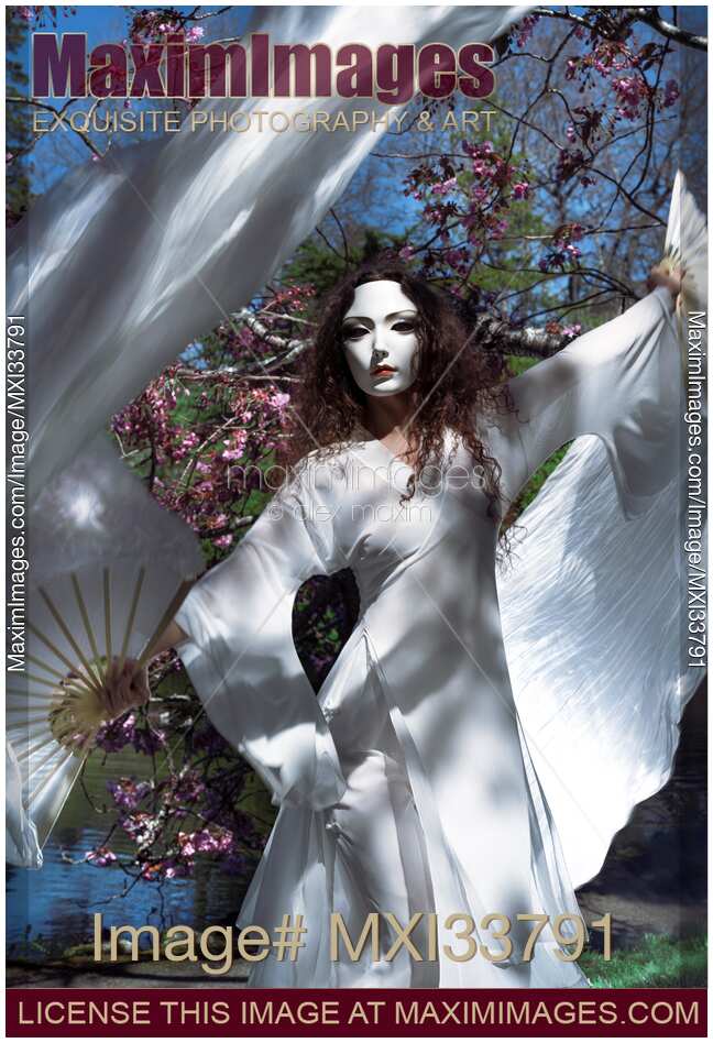 Mysterious woman in spirit-like face mask and white gown dancing gracefully with silk fans by blossoming sakura trees