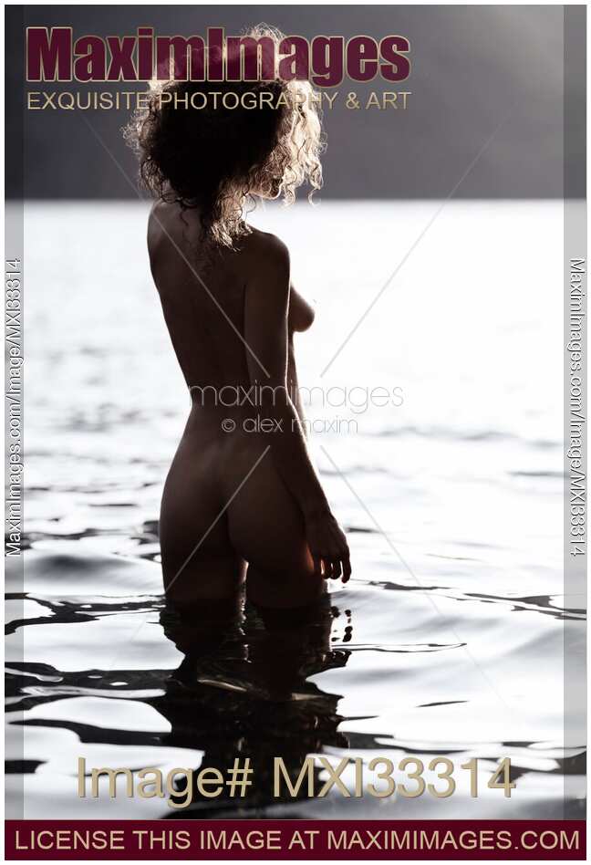 Nude woman with curly hair standing in reflective water of a lake at sunrise rear view Black and white