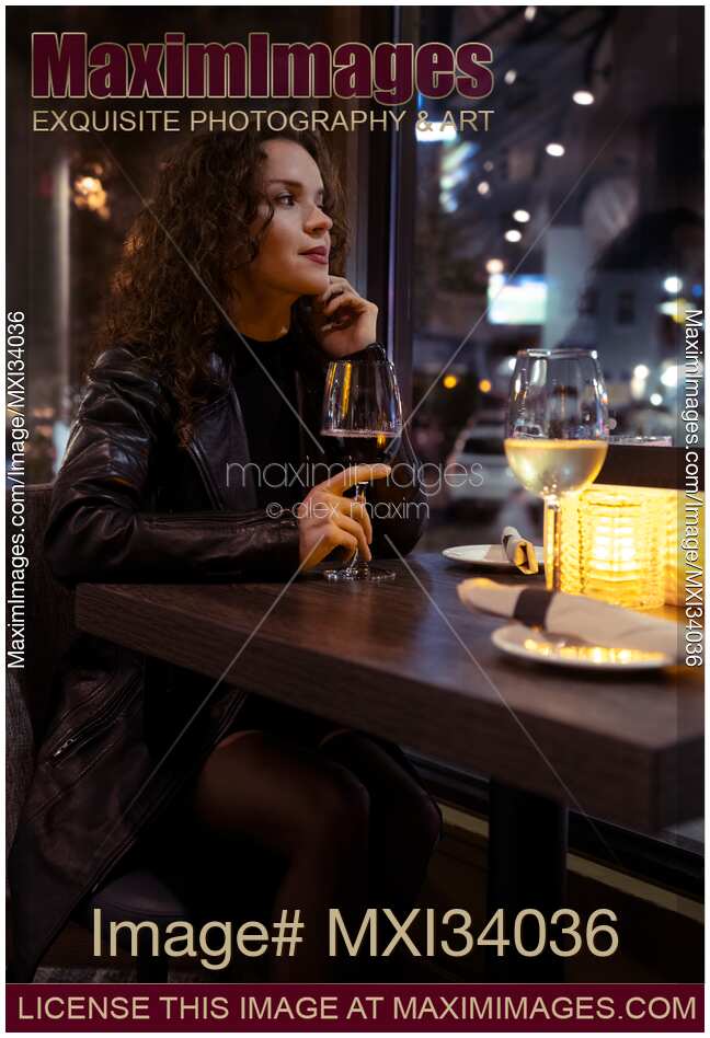 Portrait of an attractive woman in black leather coat sitting alone at a restaurant table with a glass of wine