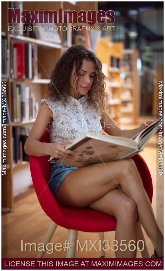 Pretty, young lady, college student reading a book in a library sitting in a chair