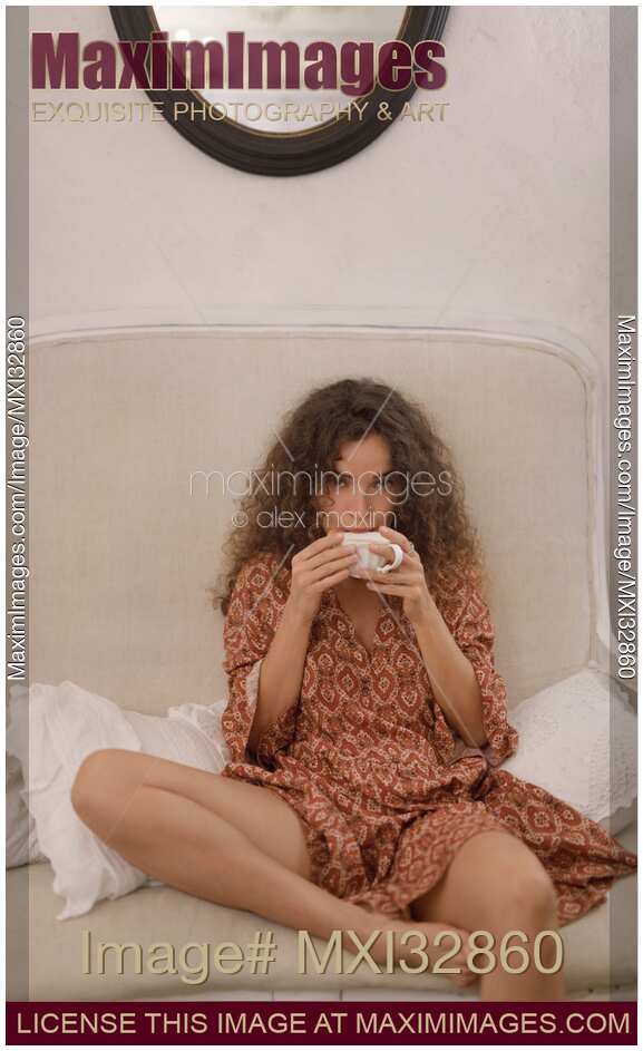 Pretty young woman in bohemian dress sitting on a white sofa sipping from a cup in a sensual provocative carefree pose