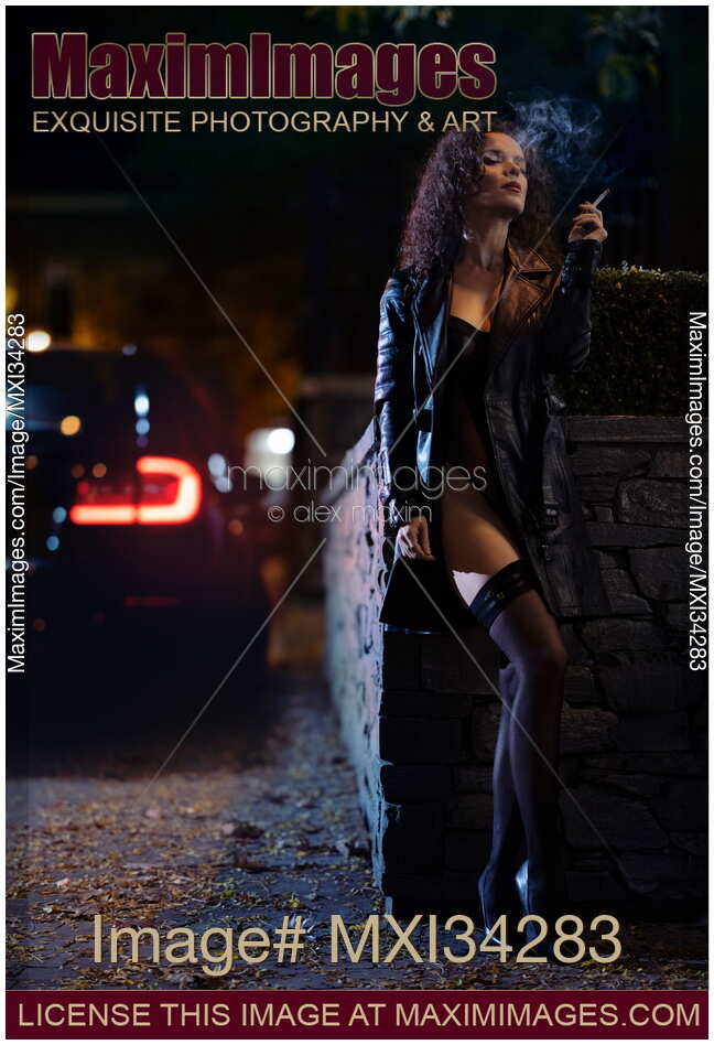 Provocatively dressed seductive woman in black leather coat smoking cigarette on night city street with car stopping by