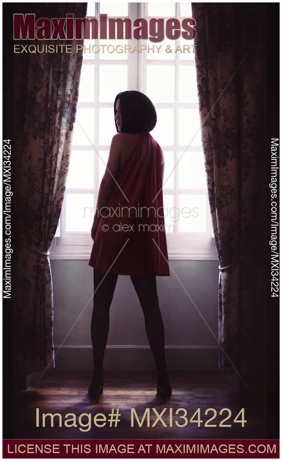 Rear view of a sexy lady in red mini dress and black stockings standing in a dark room by the window