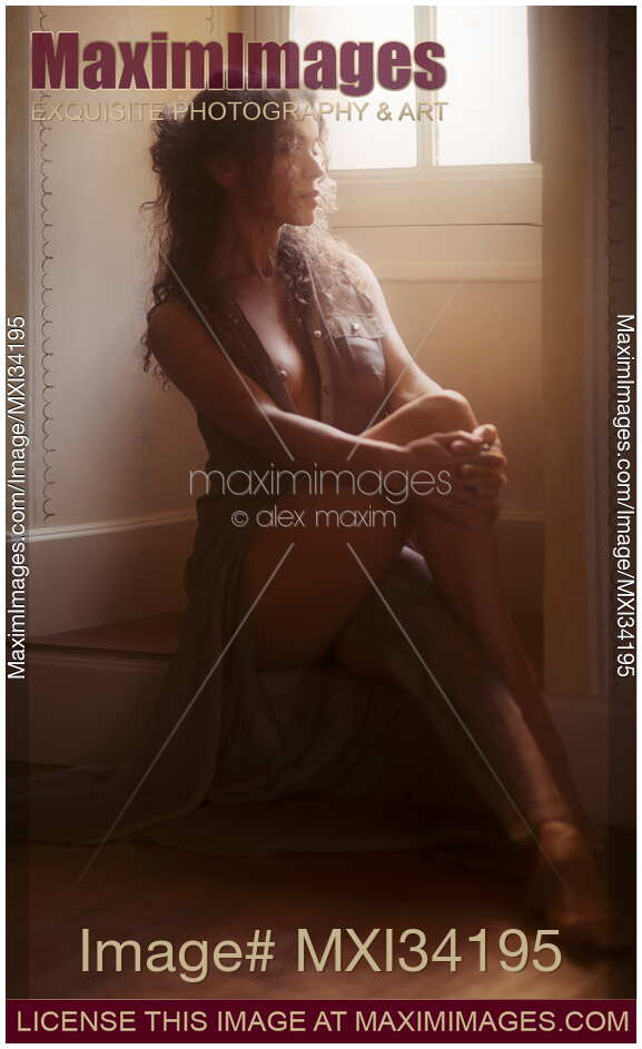 Sensual boudoir portrait of a sexy young woman in unbuttoned sheer dress sitting dreamily on the floor by a window