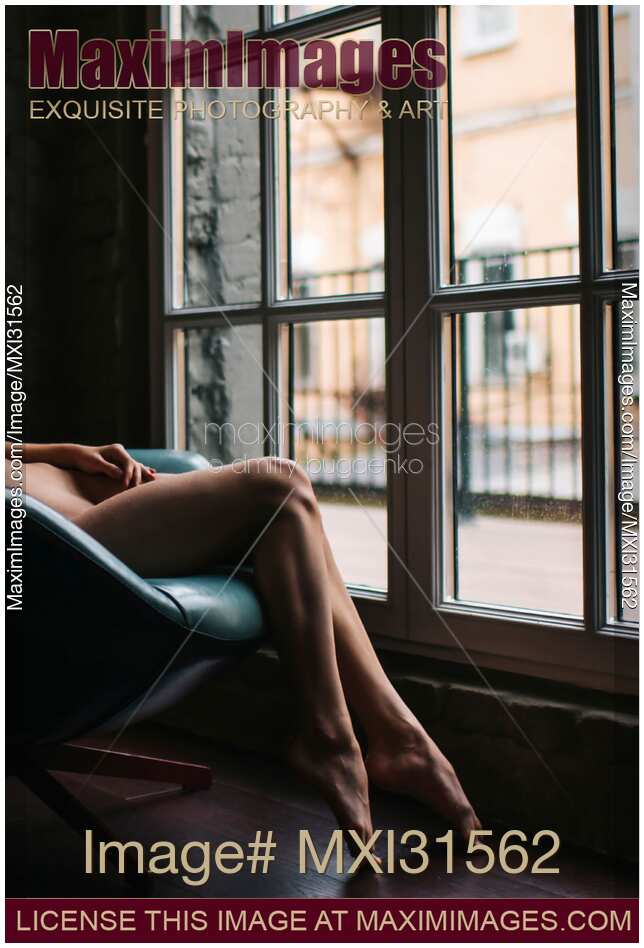 Sensual closeup of bare legs of a woman sitting naked in a chair by a large window