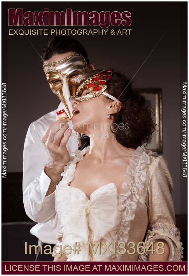 Sensual couple portrait of masked Casanova man seducing beautiful lady in vintage Venetian dress and carnival mask with cake pastry