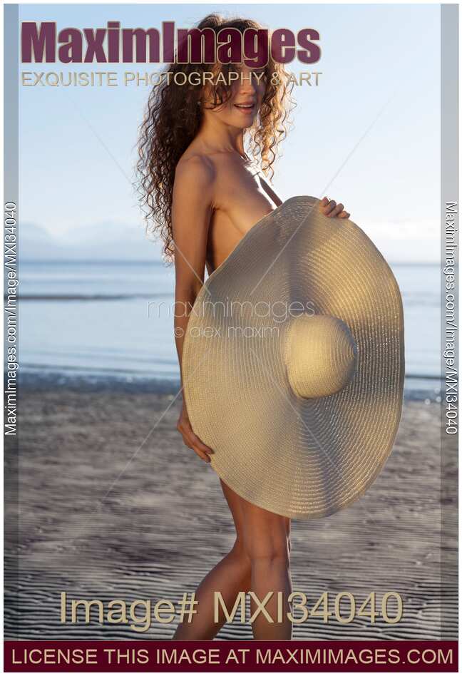 Sexy girl with a mischievous teasing look standing naked on a sunny beach seashore covering her nude body with a large sun hat