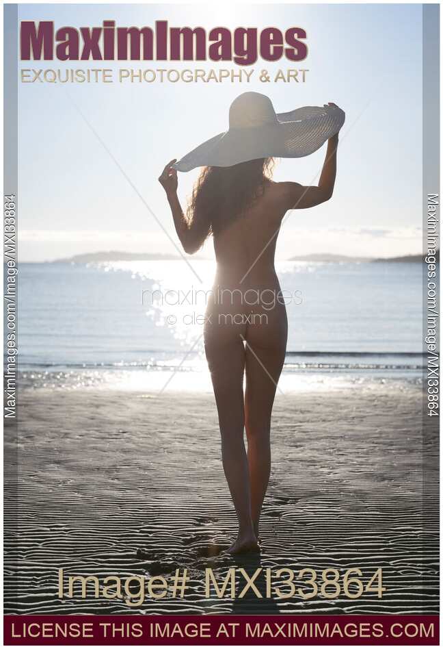 Sexy sun-tanned naked young woman in a big sun hat standing at a sandy beach in sunlight. Rear view of her nude back.