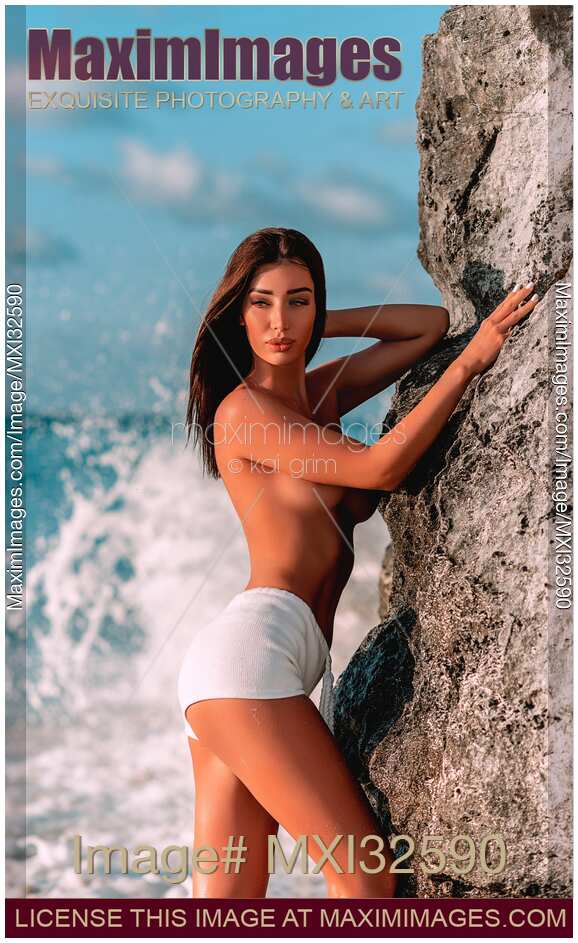 Sexy topless exotic woman at a sea shore leaning against a rock with crushing waves