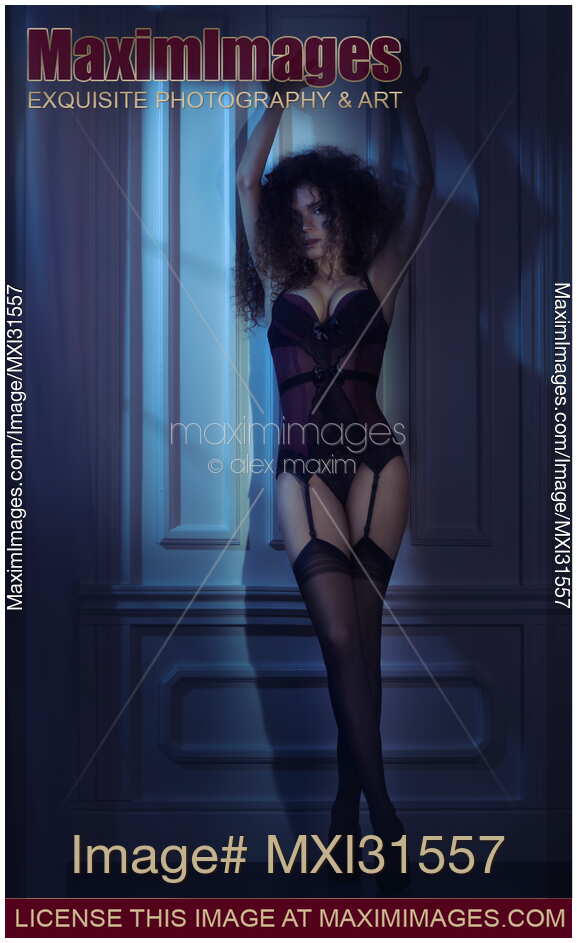 Sexy woman in a purple corset lingerie and stockings standing against the wall in dark room at night