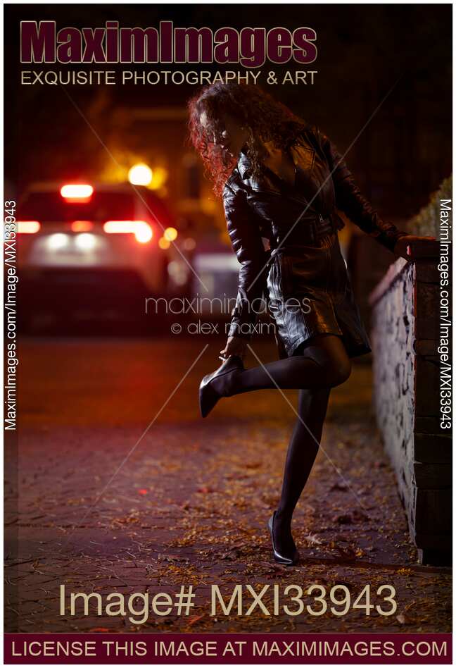 Sexy young woman in black leather coat fixing a high heel shoe on night city street in car lights
