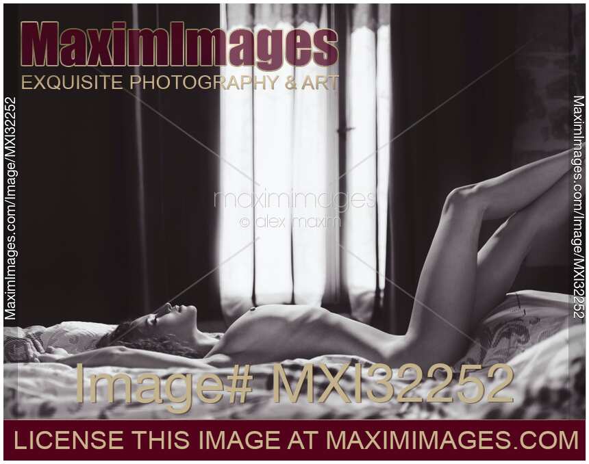 Sexy young woman lying naked stretched on a bed by a window with her legs on headboard Black and white