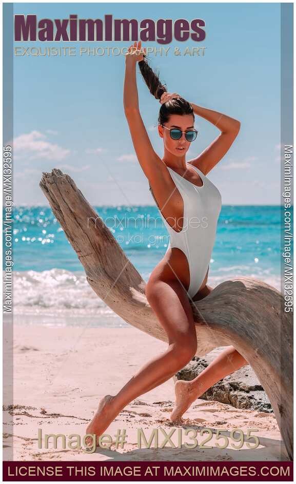 Sun-tanned exotic woman in swimsuit at a sandy sunny beach sitting on driftwood in a sexy pose Caribbean Sea