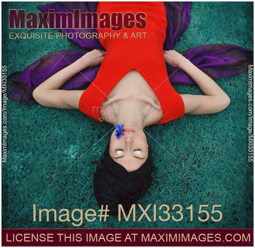 Surreal portrait of an extravagant lady in red dress lying on blue grass with a flower in her mouth
