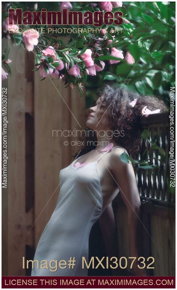 Tranquil sensual portrait of a beautiful woman under a pink camellia with fallen flower petals in her long curly hair and on her chest