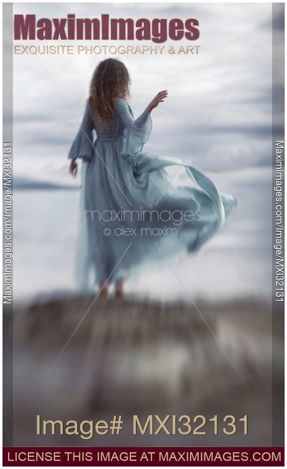 Woman in a long elegant dress flying in the wind standing on ocean shore in sunrise scenery Rear view