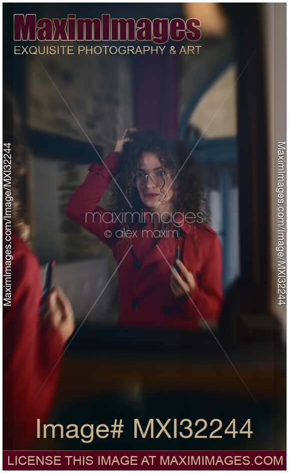 Woman in a red coat and glasses standing in front of a mirror holding red lipstick fixing makeup and hair
